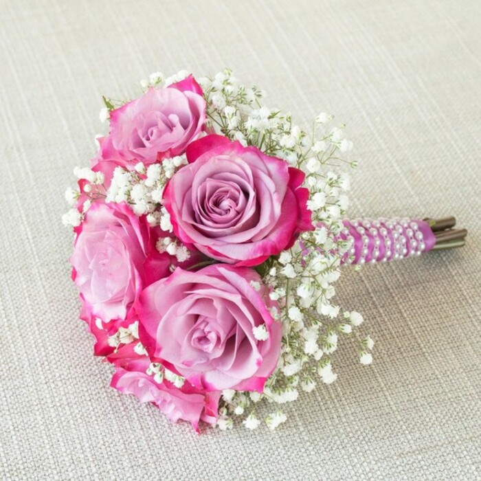 Lovingly crafted by a skilled Queensbury florist, this elegant wedding bouquet is a tribute to romance, artistry, and the gentle beauty found throughout the NW9 area. The arrangement features five prominent roses, each unfurling in a captivating blend of hues: central petals shimmer in muted lavender and softest pink, gradually shifting outward into bold fuchsia and rich magenta, creating a sense of depth reminiscent of spring sunsets in nearby Canons Park. Delicate wisps of soft white baby's breath (Gypsophila) are nestled between the roses, their frothy texture and pure tones adding volume and visual lightness, ensuring each bloom stands out brilliantly against the airy filler. The stems are trimmed short and tightly gathered, the handle encased in luscious plum satin ribbon, finished with a striking spiral of creamy white pearls-an elegant nod to tradtional bridal details favored by many couples in Queensbury. At the tip, the green-brown stems are deliberately left visible, reinforcing the bouquet's fresh-from-the-garden authenticity and bespoke character. Placed atop an oatmeal-toned, subtly textured fabric-possibly linen or fine burlap-the bouquet's vibrant colors and intricate details shine. Captured overhead, every layer and flourish speaks of meticulous craftsmanship and local pride. Perfect for a bridesmaid, bride, or as an intimate wedding centerpiece, this arrangement blends classic romance with a modern palette, perfectly complementing celebrations anywhere throughout Queensbury and the neighbouring areas.
