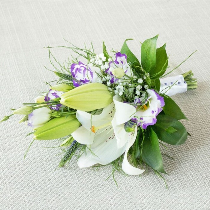 Laid diagonally across a light beige, textured fabric that resembles gently woven linen, this hand-tied bouquet of white lilies and purple lisianthus appears poised for a walk down the aisle in Queensbury. The arrangement is dominated by large, open white Oriental lilies whose petals gracefully curve outward, each bloom displaying a ring of golden-yellow to orange stamens at its centre and a delicate dusting of reddish-brown freckles across the inner surfaces. Near these fully opened flowers, several bright chartreuse-green lily buds are clustered, firm and closed, hinting that the bouquet will evolve over time as more blossoms unfurl. Interlaced throughout the design are lisianthus flowers, their ruffled, cup-shaped petals edged in rich purple that gently fades into creamy white, creating a watercolor effect of lavender and violet tones. Fine sprays of baby's breath scatter like soft white mist between the larger blooms, adding a cloud-like lightness and enhancing the impression of romance. The greenery is thoughtfully layered: broad, polished dark green leaves offer structure and a lush backdrop, while slender, wispy fronds and occasional spiky green stems extend outward, giving the bouquet movement and a slightly elongated, contemporary profile. At the narrow end, the gathered stems are wrapped neatly in a white satin ribbon that is subtly decorated with small, pearl-like beads, reinforcing the sense that this is a bridal or special-occasion piece rather than a casual bunch. The gentle, even lighting and neutral background draw the eye entirely to the flowers, highlighting the crisp white petals and purple accents. It evokes the feeling of a refined wedding or anniversary celebration at a local venue near Queensbury, the kind of elegant arrangement a trusted neighbourhood florist would prepare for same-day delivery to mark a meaningful moment.