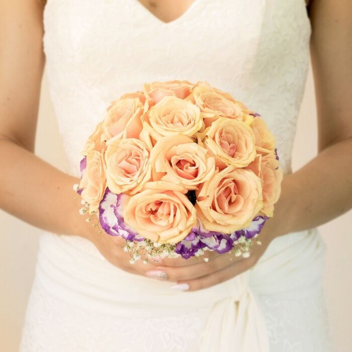 A close-up, mid-torso view of a bride in Queensbury centres on the bouquet she holds carefully in both hands, just at the level of her waist. Her gown is a classic bridal white or soft ivory, with a lace-detailed bodice and a modest V-neckline that lends refinement rather than extravagance. The fabric of the skirt falls in clean, gentle folds, catching the light in soft highlights that melt into a pale, uncluttered background, so the viewer's eye is drawn straight to the flowers. The bouquet is shaped into a perfect, rounded dome, dominated by an abundance of roses in shades of peach, light apricot, and blush pink. Each rose appears freshly opened, petals tightly layered and velvety, with the gradation of colour suggesting warmth and tenderness, like evening light settling over a quiet Queensbury side street. Beneath this rose canopy, a distinctive collar of ruffled purple blooms with crisp white edges-likely lisianthus or a similar flower-forms a rich border, giving the design both contrast and depth. Small clusters of tiny white filler flowers, reminiscent of baby's breath, are tucked thoughtfully around the base and slightly in between, adding a floating, airy feel that echoes the lace pattern on the bodice. The bride's nails are neatly manicured in a soft, understated shade, with a hint of shimmer visible on one thumb, catching a glimmer of ambient light. There are no visible accessories, no distractions-only the serene pairing of gown and bouquet. The overall atmosphere is intimate, romantic, and poised, like a moment stolen just before stepping out toward a ceremony at a local venue, the kind of bouquet that a skilled florist at Flower Delivery Queensbury might design for couples marrying close to their homes and favourite parks.
