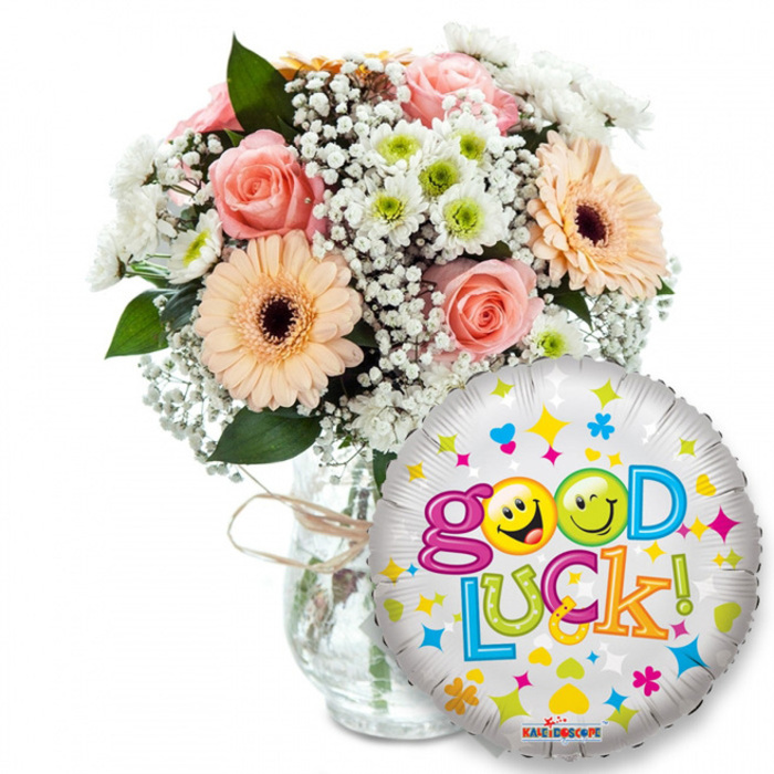 A warm, encouraging Good Luck ensemble staged against bright white: a generous bouquet sits to the left in a clear glass vase, the stems visible through the water and the neck of the vase finished with rustic twine that suggests hand-tied care. The flowers are a harmonious mix of soft peach and light pink roses with layered petals, lively peachy-orange Gerbera daisies centred in dark brown, and plenty of tiny white Gypsophila that floats like lace between the larger blooms. Small white and lime-green chrysanthemum-like florets add crisp, energetic texture while rich green foliage provides depth and contrast. Dominating the right foreground, a round metallic silver foil balloon reads Good Luck in cheerful, multicoloured lettering; two yellow smiley faces form the o's of Good - animated, playful faces that wink and grin - and the balloon is scattered with colourful hearts, stars and confetti motifs, the Kaleidoscope brand discreetly printed along the bottom edge. Soft, natural-looking studio light gives the roses a gentle glow and the balloon a reflective sheen, creating a feeling of quiet celebration. As an experienced local florist, I picture preparing this arrangement for deliveries across Queensbury - from the Broadway shops to homes near Kingsbury Road - to mark exams, job interviews, farewells or fresh starts. Flower Delivery Queensbury composes this pairing to translate hope and encouragement into a visible, fragrant gesture someone can carry with them into their next chapter.