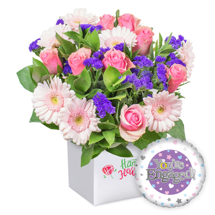 Near the heart of Queensbury, this luxurious engagement bouquet arrives in a pristine white square box, its clean, architectural lines amplifying the bouquet's sumptuous color story. Pink roses, fully opened and softly unfurling, lead the arrangement with timeless romance, while pale pink gerbera daisies lend a luminous, sunlit note with their broad petals and pale centers. Velvet clusters of purple statice punctuate the bouquet, adding depth and a regal contrast against lush green foliage that frames the arrangement like a private garden. The box, sleek and white, speaks of contemporary craftsmanship, its surface subtly decorated with a refined red rose motif rather than explicit branding, signaling a heartfelt, local touch from a Queensbury florist. A round foil balloon anchors the composition in the lower right, its face a central purple circle framed by a white ring dotted with pastel hearts and silver stars, the words You're Engaged! gleaming in silver. This is a refined gift suitable for a refined home or a romantic toast at a Kingsbury venue after a proposal, and it is available for dependable same-day delivery across Queensbury, Kingsbury, and Burnt Oak through our local service. Crafted with care, the bouquet promises warmth, joy, and enduring romance for the couple's bright future together.