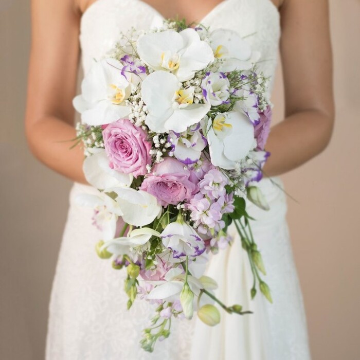 In this evocative, close-up composition, a Queensbury bride-seen from the chest down-holds a grand, cascading bridal bouquet, the image suffused with a radiant softness provided by a gently blurred, light beige background. Her strapless white gown, featuring intricate lacework at the bodice and upper skirt, enhances the scene's sense of sophistication. The bouquet itself is a study in harmony: prominent, snowy white Phalaenopsis orchids with buttery yellow and subtle purple centres, their petals broad and serene, are paired with romantic blush-pink garden roses luxuriously in full bloom. Soft clusters of lavender and light purple stock, with their marshmallow-like spiked texture, thread through the lush arrangement, joined by hints of white lisianthus-some edged in deep purple-providing an extra layer of detail. Wisps of baby's breath and delicate green foliage are woven throughout, creating a natural, elongated cascade. The overall effect is one of delicate opulence and timeless beauty, conjuring thoughts of weddings near Kingsbury Circle or intimate ceremonies in Queensbury's leafy venues. The image beautifully distills the artistry and care typical of local florists, evoking both quiet anticipation and enduring romance for couples tying the knot in this vibrant North London enclave.