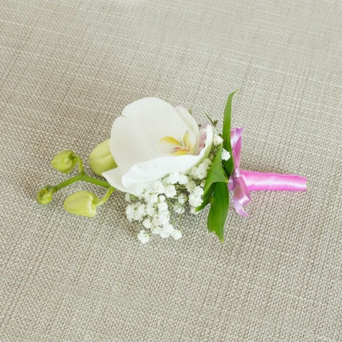 An elegant orchid boutonniere is displayed horizontally on a neutral, light beige linen-style fabric, its understated background allowing every floral detail to stand out with clarity and grace. At the centre sits a large, pristine white Phalaenopsis orchid, its broad petals gently arcing outward in smooth, velvety curves. The flower's throat shows delicate accents of soft yellow and the most subtle blush of pale pink, like a gentle sunrise caught within the bloom, offering warmth without overpowering the composition. Clustering around the base of the orchid are many tiny white sprigs of baby's breath, each miniature flower forming part of a soft, cloud-like ring that adds volume and a dreamy, ethereal texture. Just behind, a few slender, bright green leaves offer a fresh, clean contrast to the white and pastel tones, suggesting a living connection to the garden, much like the greenery found in the quieter corners of Queensbury's residential gardens. To one side, a narrow stem extends outward, carrying several tightly closed, lime-green orchid buds that hint at future blossoms and add a sense of natural growth and unfolding stories-ideal for symbolising new beginnings at weddings and milestone celebrations. The stems of the entire arrangement are carefully gathered and secured with a narrow, luminous fuchsia pink satin ribbon, tied into a precise, elegant bow with short trailing ends. The ribbon's glossy surface catches the light, injecting a sophisticated splash of colour that feels festive yet refined, perfect for a groom, groomsman, or prom guest preparing for an event in or around Queensbury. Gentle, even lighting washes over the scene, revealing the fine weave of the fabric, the smooth orchid petals, and the airy tufts of baby's breath, creating a calm, polished mood that speaks of thoughtful floral design and quiet celebration.