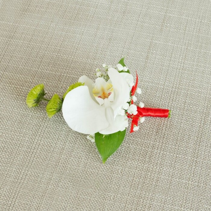 On a softly textured light beige linen surface, this refined boutonniere is captured in a moment of stillness, as if waiting to be pinned to a lapel before a ceremony at a nearby Queensbury church or registry office. Dominating the small composition is a single, open white Phalaenopsis orchid, its broad petals smooth and luminous, curving outwards in a graceful, almost sculptural form. In the centre of the bloom, a soft yellow-gold throat gently melts into flecks of reddish-orange or warm brown, the natural markings subtle yet intricate, giving the impression of fine brushstrokes on porcelain. Surrounding the orchid, airy stems of white baby's breath have been tucked in with care, their clusters of tiny, cloud-like blossoms adding a whisper-light texture and a sense of movement, as though a delicate breeze might stir them. To the left, two round lime-green chrysanthemum button blooms, with their compact yet slightly spiky petals, introduce a lively burst of green that feels both modern and cheerful, echoing the fresh tones of Queensbury gardens in early summer. A deep green leaf fans out at the base of the orchid, providing a glossy, natural foundation, while a smaller, darker leaf peeps from near the stem, adding layered dimension to the greenery. The stems are tightly bound in a striking crimson red satin ribbon, which is neatly wrapped and finished in a small bow or knot, creating a polished handle for the florist's work and offering a theatrical contrast to the calm whites and greens. The linen-like fabric beneath, with its slightly coarse weave and neutral tone, underscores the boutonniere's elegance without competing for attention, letting every hue and texture stand out clearly. This floral pin feels perfectly suited to a groom, groomsman, or stylish guest, blending classic wedding tradition with a touch of contemporary colour that reflects the thoughtful craftsmanship of local florists serving the Queensbury community.