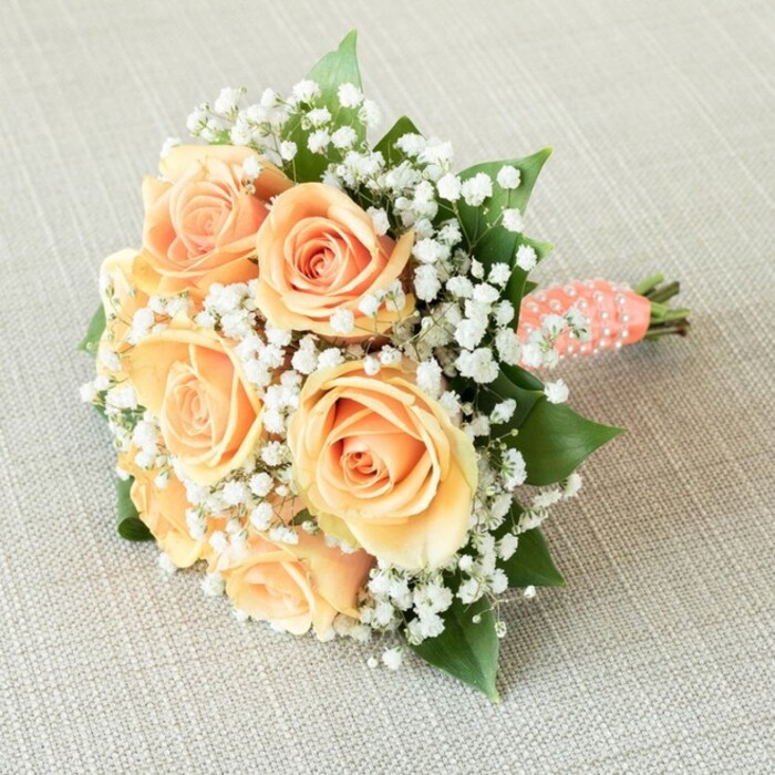 A vision of elegance and softness, this round bridal bouquet is artfully arranged and displayed against a finely textured, oatmeal-beige linen reminiscent of the inviting table settings found throughout Queensbury's cherished family homes. Seven radiant roses bloom in perfect harmony at the bouquet's center, their petals unfurling gracefully in gradients of soft peach, blushing apricot, and delicate pale orange, each catching the soft diffuse light in a gentle embrace. Interlaced among these focal roses are delicate clouds of baby's breath, or gypsophila, their tiny white flowers adding depth and an ethereal sense of airiness that gently contrasts with the warmth of the roses. Hints of dark green, glossy rose leaves peek from beneath the arrangement, grounding it with a touch of organic freshness often found in Queensbury's green spaces. The beautifully gathered stems are wrapped in a blush-coral satin ribbon, which is elegantly finished with a spiral of tiny white faux pearls-details beloved by brides seeking timeless beauty with a touch of Queensbury charm. The bouquet's base, where the stems are neatly cut and exposed, adds a note of authenticity. The entire composition radiates under soft, ambient light, highlighting the artistry and care that local florists bring to each sudden celebration-from intimate ceremonies in Roe Green to anniversary dinners along Kenton Road. This bouquet embodies romance, sophistication, and the enduring style of Queensbury's floral traditions, making it the perfect choice for weddings, showers, and all special moments.
