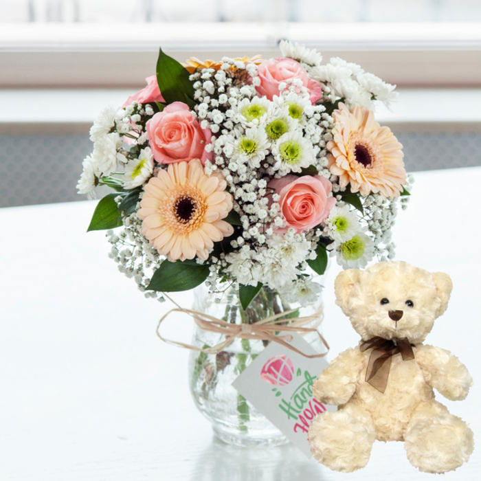 A gentle, love-filled still life is arranged on a crisp white surface, the kind of scene you might find on a carefully set table in a Queensbury flat ready for an anniversary surprise. In the centre, a rounded bouquet sits in a clear glass jar-style vase, its transparency showing the fresh green stems resting in clear water. The floral mix is beautifully balanced: soft pink roses with curving, layered petals; pastel peach gerbera daisies whose deep dark brown centres draw the eye; delicate tufts of white baby's breath that soften the composition; and small chrysanthemum blooms in clean white, some with a hint of lime-green at the core to add a modern freshness. Around the vase neck, natural jute-style twine is tied simply, holding a small white rectangular tag. On the tag, a pink heart graphic and the word Handmade in graceful green script quietly suggest that this gift has been crafted with intention rather than mass-produced. To the right, a plush cream teddy bear sits upright, angled slightly toward the arrangement as though keeping it company. Its fur appears invitingly soft, with tiny dark eyes that catch the light, a matching brown nose, and a slim dark brown satin ribbon secured neatly in a bow around its neck. The faint reflection of both bouquet and bear on the polished white surface adds a subtle sense of depth, while the background blurs into bright, airy tones that feel like daylight streaming in from a window near Queensbury's shops or local cafés. The overall mood is warm, intimate, and celebratory, making this duo ideal for anniversary gifts, romantic gestures, or heartfelt same-day flower delivery in Queensbury.