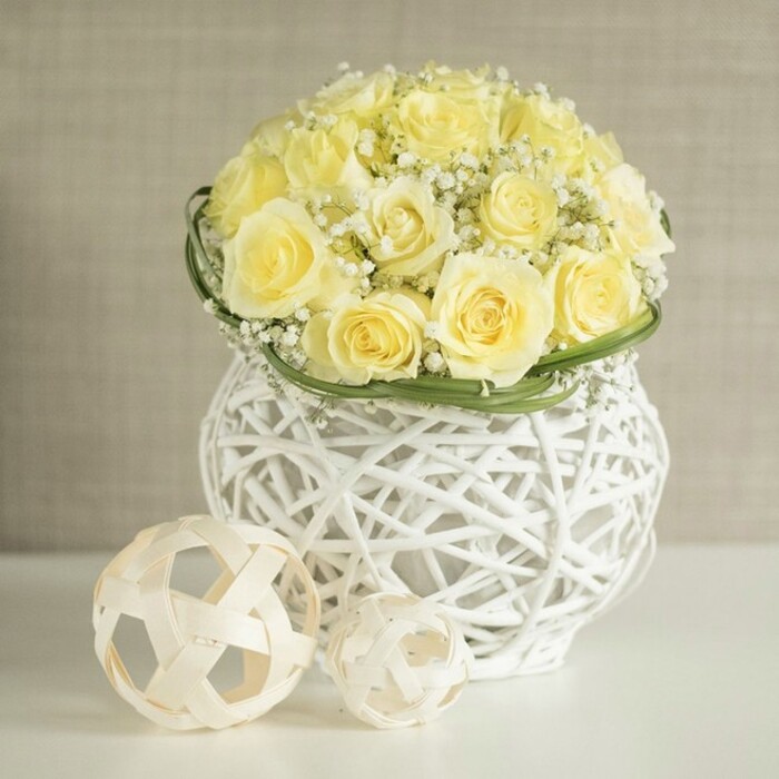 A refined floral centerpiece is carefully arranged as though waiting on a wedding table in Queensbury, perhaps in a light-filled dining room overlooking the quiet residential streets. Dominating the composition is a perfectly rounded dome of pale yellow roses, each stem positioned with care so that the blooms touch and overlap, forming a continuous surface of velvety petals. The colour sits between cream and soft butter, warm yet gentle, evoking the calm of a late spring afternoon. Interwoven among these roses are numerous sprigs of white baby's breath, their minuscule blossoms forming frothy clusters that nestle into the gaps and soften the structure, adding delicate volume and a sense of dreamy, airy movement.
Encircling the base of the bouquet is a slim ring of green foliage, likely flat, pliable leaves that have been wrapped to create a clean, polished finish. This touch of vivid green neatly separates the mass of flowers from the vessel beneath, acting like a natural ribbon. The bouquet is presented in an eye-catching white vase fashioned from an intricate woven framework, similar to finely crafted wicker or rattan but with a contemporary twist. Its rounded shape, slightly narrower through the middle, gives the arrangement an elegant silhouette, while the open weave allows light to play through, sending subtle shadows across the smooth white tabletop.
On the table, two decorative spheres, also made from slender, pale wooden strands, sit close to the vase. Their irregular geometric weave reflects the pattern of the container, reinforcing the theme of natural textures and handcrafted detail. The softly blurred, neutral-toned background suggests a calm interior typical of Queensbury homes, where beige and cream walls make a perfect canvas for floral displays. Together, the roses, baby's breath, and woven accents convey a mood of quiet celebration and sophisticated simplicity, ideal for wedding receptions, anniversary dinners, or thoughtful styling for local Queensbury events.