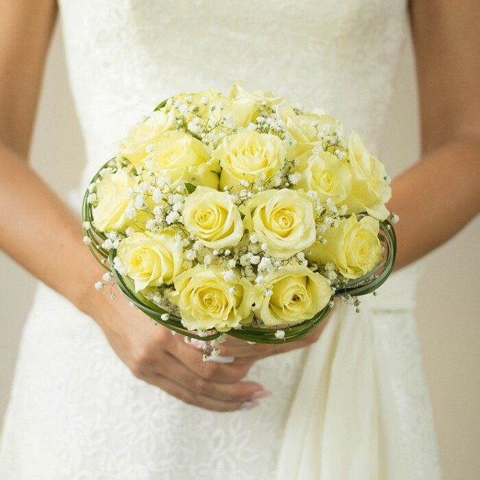 This close-up reveals a bouquet that epitomises the joyful spirit of a Queensbury wedding, lovingly held at the centre of a scene rich with anticipation. The main feature is a tightly packed, rounded display of soft yellow roses-each blossom perfectly formed, their delicate petals catching and diffusing the gentle light filtering in from a local celebration space. Wispy sprays of pure white baby's breath are artfully tucked among the roses, lending the arrangement a touch of ethereal lightness and evoking the delicate spring mornings often enjoyed along Honeypot Lane. At the base, thin bands of dark green foliage or fine decorative wire circle the bouquet in graceful loops, enhancing its geometry and reinforcing its sense of refined handmade quality. The hands that cradle this creation-a gentle curve beneath the blooms, nails neatly manicured in natural tones, and a simple, radiant ring-speak to the personal significance of the moment. The bride's dress, glimpsed in a backdrop of soft, textured white, hints at intricate lacework quietly framing the flowers. The entire composition is gently blurred around the edges, ensuring that the gentle luminosity and complexity of the bouquet are the stars of the photograph. In every sense, this image tells a story of lasting romance, the artistry of Queensbury florists, and the classic beauty suited for the most cherished of celebrations.