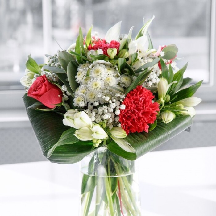 This image presents a beautifully balanced red and white bouquet, styled with the precision and warmth you might expect from a long-established Queensbury florist. The flowers sit in a straight-sided, clear glass vase filled with fresh water, through which the trimmed green stems appear almost architectural, forming a strong, vertical base. Above, a generous dome of blooms unfolds. Deep, velvety red roses with gently unfurling petals mingle with a bold, ruffled crimson carnation at the front, the carnation's many layers creating fascinating shadows and a rich, tactile depth. These passionate reds are offset by soft whites and creams: elegant white alstroemeria with slender, speckled petals, rounded white chrysanthemums or similar blooms, and several closed white lily buds whose stillness adds a quiet, poised energy, as though the bouquet is mid-story and more beauty is yet to come. Sprigs of gypsophila-tiny white baby's breath-thread through the composition, their cloud-like clusters lending an ethereal, celebratory feel, much like confetti at a local Queensbury gathering. The foliage is equally considered: large, glossy dark green leaves are curved and wrapped around the base, forming a polished collar that frames the flowers, while pointed, eucalyptus-like greenery adds texture and soft silvery-green tones higher in the arrangement. Everything rests on a spotless white surface, set against a bright, softly blurred background suggestive of daylight streaming through a window in a Queensbury home or office. The lighting is gentle and even, enhancing each colour and curve without harshness, conveying an atmosphere of calm joy and understated sophistication-a centrepiece suited to anniversaries, birthdays, or simply brightening an ordinary day with a touch of local floral artistry.