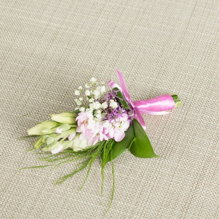 Centred on a softly textured, neutral linen surface, this beautifully composed boutonniere or small corsage evokes the quiet elegance of a Queensbury celebration, whether it's a garden reception near Roe Green or an intimate dinner on one of the tree-lined residential streets. The fabric beneath is a pale oatmeal or light greige hue, its subtle weave creating a gentle backdrop that lets every floral detail stand out. At the core of the arrangement is a cluster of soft light pink blooms, their many petals finely ruffled and gathered close together, calling to mind miniature stocks or dainty garden sprays. The petals display a tender gradient, with slightly deeper pink tones nestled toward the centres and lighter, almost pastel tips, giving the impression of petals just brushed by morning light. Threaded throughout are tiny white baby's breath blossoms, forming airy tufts that lend the piece a cloud-like, romantic softness. Delicate accents of lavender or light purple florets peek out between the pink petals, adding a hint of cool colour and visual texture, much like the subtle pops of floral colour you might see in front gardens walking through Queensbury. To one side, a row of elongated, tightly furled buds in creamy white and pale green-similar to young freesias or slender lilies-rests gracefully, their closed forms suggesting freshness and new beginnings. Supporting the flowers is a base of lush, broad green leaves, rich and vibrant, while whisper-fine, grass-like or ferny green sprigs extend beyond the main cluster, creating a soft, slightly untamed edge. The stems are cleanly gathered and wrapped in a bright fuchsia satin ribbon, the material smooth and lustrous, tied into a simple yet refined bow whose loops and short tails fall neatly along the fabric. The overall impression is one of careful craftsmanship and understated celebration, ideal for a groom's lapel, a prom corsage, or a special guest's accessory prepared by a skilled Queensbury florist.