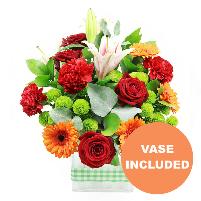 An exuberant mixed bouquet is captured in sharp detail against a bright white background, the colours appearing almost jewel-like in the clear, diffused light. The flowers are arranged in a sturdy, rectangular clear glass vase, where the straight, carefully trimmed stems can be seen through the water, suggesting professional florist care and freshness ideal for same-day delivery around Queensbury. Wrapped snugly around the lower part of the vase is a light green and white gingham band, creating an inviting, country-style accent that would look at home on a kitchen table overlooking the quiet residential streets near Kingsbury High Road. The front of the bouquet is dominated by fully opened deep red roses, their velvety petals spiralling inward, flanked by similarly rich red and red-orange carnations whose frilled edges lend a soft, ruffled texture. Scattered between them, bright orange gerbera daisies with dark, almost black centres tilt their large, flat faces outward, giving the arrangement a sense of cheerful animation. Punctuating this warmth are vivid lime-green pompom chrysanthemums, their small, perfectly round blooms tucked into gaps to create visual rhythm and a playful, contemporary twist. Rising above the rest, tall white Oriental lilies bring elegance and height; some trumpets are wide open, revealing pale pink stripes and prominent stamens, while others remain in smooth buds that will extend the life and changing look of the display over several days. Threads of glossy deep green foliage cradle the blooms, while long, slender stems of silver-green eucalyptus arch gracefully from the sides, adding movement and a soft, misty tone to balance the stronger colours. In the lower right corner of the image, a solid orange circular badge overlaps a portion of the vase, with the clear white text VASE INCLUDED announcing that this arrangement arrives as a complete, ready-to-place gift.