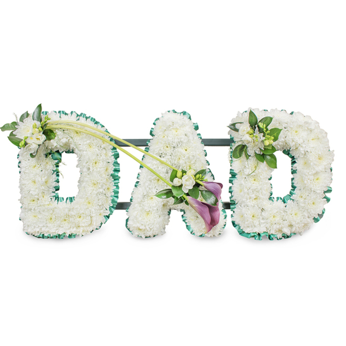 A beautifully composed funeral tribute spells out DAD in large block letters, the kind of design that might rest beside a coffin during a Queensbury service or be placed by the graveside at the end of a quiet farewell. Each letter is formed from countless white chrysanthemums, packed so tightly that they create a flawless, quilted surface of petals. The blooms are crisp and freshly opened, their soft texture and snowy colour evoking innocence, respect, and a gentle stillness. Around each letter, a deep emerald green ribbon is folded into neat ruffles, tracing every curve and straight line, offering a vivid contrast that frames the tribute and hints at the greenery of nearby Queensbury Park or the tree-lined streets around it. The first D carries a tasteful accent on its upper left edge: a small spray of white freesia and polished green leaves, the buds and open flowers adding fragrance and an airy lightness. From this point, two long, arching green stems sweep diagonally across towards the A, giving the whole arrangement a sense of graceful movement. At their meeting point at the lower right of the A, an eye-catching spray creates the emotional heart of the piece. Here, two deep purple calla lilies, with their sleek, trumpet-shaped blooms, are nestled among yet more white freesia and rich foliage, their darker colour symbolising depth of feeling and solemn devotion. The final D is gently embellished on its upper right with another modest cluster of freesia and greenery, echoing the first letter. Seen against a clean white background, this DAD tribute feels deeply personal and thoughtfully arranged, a loving expression of remembrance for a father cherished by his Queensbury family.