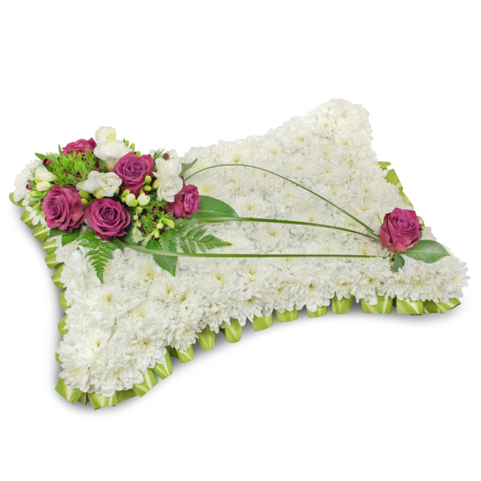 A classic pillow-shaped funeral arrangement rests on a stark white background, the simple setting emphasizing the thoughtful construction and peaceful mood. The main surface is completely covered in hundreds of tiny white chrysanthemum florets; their tight, repeated forms create a soft, cloud-like tapestry that suggests rest and purity. Encircling the cushion, a neat trim of light-green satin ribbon has been folded into a subtle ruffle, offering a refined edge that brings a hint of colour and a tailored finish. In the top-left corner a concentrated cluster of deep magenta roses commands attention-their velvety petals opening in rich, saturated layers-while small white freesia blooms nestle between them, introducing delicate points of brightness and a gentle citrus-tinged scent. Lush green foliage, including airy fern fronds and broader leaves, provides a verdant backdrop that gives the cluster volume and contrast against the white chrysanthemum field. Across the face of the cushion, three slender bear-grass stems sweep in elegant arcs from the floral cluster toward the bottom-right, imparting a refined, calligraphic line to the design; a single magenta rose sits near that lower corner, placed with intention to counterbalance and complete the composition. The lighting is soft and even, catching slight sheen on satin ribbon and the velvety texture of rose petals, creating an atmosphere of gentle reverence. This carefully composed pillow, suitable for memorial services across Queensbury and nearby Kenton, communicates comfort, respect and enduring remembrance.