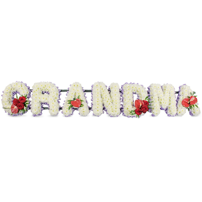 A lovingly crafted floral tribute spells out the word GRANDMA in striking block capitals, stretching horizontally across the frame against a simple white background that keeps every detail clear. Each letter is densely packed with pristine white cushion chrysanthemums or similar small-headed blooms, creating a smooth, velvety surface that looks almost like fresh snow. The repetition of the tiny petals gives a soft, tactile impression, as though you could run your hand over the letters and feel nothing but gentle texture. Along the edge of each character, a delicate band of soft lilac or pale purple ribbon provides a subtle outline, bringing a whisper of colour and giving depth to the design. The emotional heart of the piece lies in the carefully placed red accents: at the base of the letter G, a cluster of deep red roses is nestled beside a distinctive variegated red-and-white calla lily, its sculpted, flaring shape catching the eye. The first A is graced with a single heart-shaped red anthurium, its glossy surface reflecting the light. The central M carries another spray of rich red roses intertwined with greenery and a further red anthurium, while the final A is completed with one more anthurium bloom, echoing the earlier touches and tying the tribute together visually. Beneath and behind the letters, a dark green supporting frame holds everything neatly in line, largely hidden but giving structure and stability. The arrangement radiates calm respect and intimate love, like something a family in Queensbury might have delivered from Flower Delivery Queensbury to St. Andrew's Church or a local crematorium to say a deeply personal goodbye to their grandmother.