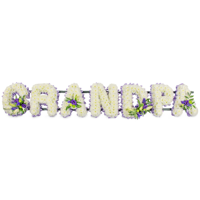 A poignant floral lettering display spells out the word GRANDPA in generous, upright capital letters, carefully arranged in a single row against a plain white background, reminiscent of how it might be presented at a funeral service in Queensbury. Each letter is entirely carpeted in small white chrysanthemums or carnations, giving the impression of soft, rounded cushions of petals. The flowers are tightly massed, creating a smooth, even texture with a gentle sheen where the light catches the curved petals, suggesting freshness and the faint, clean scent typical of white funeral blooms. Among the white, clusters of rich purple and soft lavender flowers, perhaps lisianthus or miniature chrysanthemums, are artfully tucked into selected letters, accompanied by glossy green foliage. These accents appear in the cradle of the 'G', in the centre of the first 'A', through the upright of the 'D', around the bowl of the 'P', and enhancing the final 'A', adding rhythm and visual interest to the word. A dark green bar or frame runs along the underside of the letters, largely concealed but offering stability so the tribute could sit securely on trestles beside the coffin or on a stand at a local crematorium near Queensbury. The design has the calm, respectful presence of a piece crafted with great care, like something a family would order from Flower Delivery Queensbury when they wish to honour a devoted grandfather. The soft contrast of white and purple conveys reverence, remembrance, and a gentle sense of comfort, making this tribute a meaningful focal point during moments of reflection and farewell.