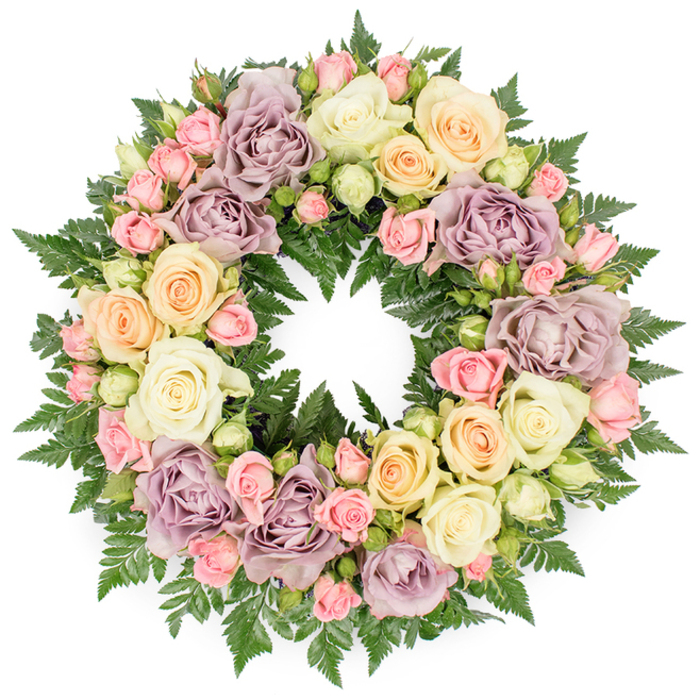 Set against a bright white background, a lush circular funeral wreath of roses and fern foliage offers a calm, dignified presence, much like the thoughtful tributes created for memorial services around Queensbury and the surrounding streets. The wreath is generously filled, creating an opulent ring of petals and greenery that appears soft yet substantial. Large, fully opened roses in creamy white and pale ivory anchor the design, their petals layered and slightly cupped, forming gentle spirals that draw the eye inward. Nestled between them are clusters of delicate blush pink spray roses; some are still in tight bud, while others are just beginning to soften and open, adding fine detail and a sense of tender progression. Soft peach and buttery yellow roses weave warmth into the palette, while distinctive dusty lavender and muted mauve roses introduce an antique, romantic nuance that feels both nostalgic and comforting. All of these blooms are supported by a thick, verdant bed of dark green fern fronds, their finely divided leaves peeking out between flowers to create depth and a lively contrast. The carefully balanced colours move from cool ivory to warm peach and back to subtle lavender, giving the wreath a harmonious, painterly quality. Light falls evenly across the arrangement, illuminating each curve and fold of the petals without glare. The circular shape, uninterrupted and perfectly formed, evokes eternal love and remembrance, making this wreath an especially meaningful choice for honouring a loved one in local chapels around Queensbury or nearby Edgware. Its overall mood is serene and consoling, a floral embrace crafted to offer quiet support in times of loss.