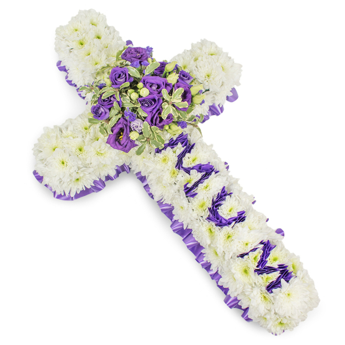 A substantial, handcrafted sympathy cross composed of tightly packed white chrysanthemums, shown against a neutral white backdrop that emphasises texture and form. The chrysanthemums create a seamless, cushiony plane across the entire face of the cross, evoking purity and calm. Encircling the cross's edge is a ruffled band of deep purple satin ribbon, neatly pleated to form an elegant border that speaks of solemnity and respect. The vertical section bears the name MUM arranged in clear, stylised lettering made from smaller purple blooms-likely eustoma or spray chrysanthemums-each petal selected for consistent hue so the message reads with dignity against the white cushion. At the arm junction a lush, compact spray commands attention: deep violet roses with velvety petals, lighter lavender lisianthus or eustoma with ruffled edges, and variegated foliage whose lighter margins and darker centres provide contrast and depth. Small clustered green berries and slender stems add texture and a hint of natural movement. The cool palette of whites and purples, combined with the satin's soft sheen, creates a composed, reverent mood appropriate for funerals, memorials or church services in Queensbury and surrounding neighbourhoods such as Colindale and Kingsbury. As a local florist I imagine this piece positioned beside a casket or on an easel at a chapel in the area, a crafted gesture of enduring affection and respect.