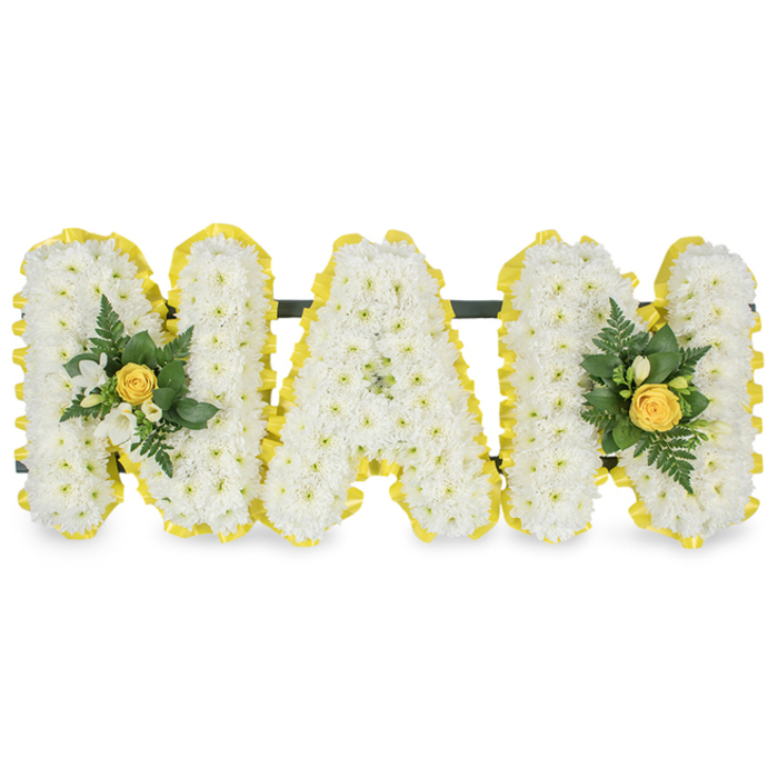 A vibrant, lovingly hand-crafted floral tribute spells the affectionate word 'MAM' in three freestanding capital letters, each letter densely covered in small, fresh white chrysanthemum sprays to produce a tactile, intricately textured surface. The perimeter of every letter is neatly edged with a bright, sunny yellow satin or pleated ribbon that provides a warm, optimistic frame and a clear visual outline from a distance. Two floral embellishments are placed with careful symmetry: one nestles against the left upright of the first 'M' and the other mirrors it on the right upright of the second 'M'; each accent centers on a soft yellow rose bud, surrounded by tiny white freesia-like blooms and an assortment of deep green foliage-slender fern fronds and glossy leaves-that give the clusters a verdant, living contrast to the white letters. A slim, dark support bar is discreetly fixed behind the upper edges of the letters to hold the piece together and allow it to be displayed upright at a graveside or memorial setting. The composition sits against a pure white background, where studio light picks up the gentle shadows between chrysanthemum heads and the satin sheen of the ribbon. This arrangement-classic white with cheerful yellow highlights-is intended as a personal, heartfelt homage to a mother figure, suitable for placement in a chapel or church near Queensbury or delivered to services across the local area, carrying with it a message of purity, warmth, and enduring affection.