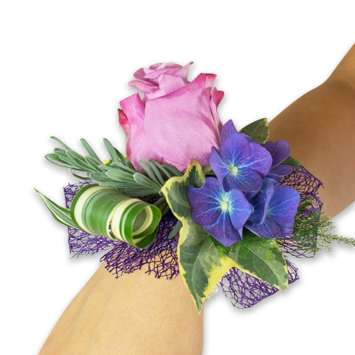 Displayed along a light-toned forearm with a stark white background that heightens every hue, this wrist corsage is a study in contrast and careful proportion. At its heart a partially open rose in a soft pink-lavender tone shows layered, velvety petals whose ruffled edges carry a deeper magenta blush, creating an elegant focal point. Flanking the rose are dense clusters of indigo-purple hydrangea; each minuscule floret reveals a pale blue-white center that seems to glow from within, adding intricate texture and a cool counterpoint to the rose's warmth. Glossy, variegated ivy leaves-dark green at the center with cream-tinted margins-spill outward, offering a formal cadence and a touch of classic garden charm. Interspersed are long, thin silvery-green sprigs, likely eucalyptus or a lavender cousin, that provide a soft, fragrant silhouette and a whisper of movement. A contemporary accent appears as a broad green leaf striped with white, curled into a tight spiral that reads like a botanical ribbon and brings sculptural interest. Underlying the arrangement, open-weave mesh in a rich violet provides a textile base-almost like a shadowed petticoat-that amplifies the purple notes and anchors the composition visually. The corsage is crafted to sit diagonally so the main bloom faces outward when worn; the textures invite a tactile glance-the silky rose, the papery hydrangea, the smooth ivy-and the lighting favours soft highlights across petals and leaf veins. Ideal for formal dances, weddings, or celebratory evenings, the piece carries a timeless yet modern sensibility suited to special moments in Queensbury's community.