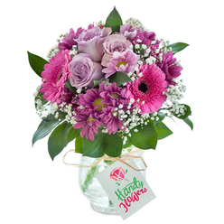 Radiating warmth and affection, this bold floral bouquet, carefully crafted by Queensbury florists, stands out in a rounded display against a bright, clean canvas. At its core, pale lavender roses exude grace, their unfolding petals gentle as a dawn breeze drifting through the gardens near Queensbury Station. Encircling the roses, brilliant fuchsia Gerbera daisies create a lively, jubilant focus, their large faces inviting admiration and symbolising cheerful wishes. Deeper magenta, plum-hued chrysanthemums interweave with sprays of delicate white baby's breath, adding layers and a cloud-like softness, while lush greenery escapes the cluster, providing freshness and a natural flow akin to the leafy streets around Honeypot Lane. The arrangement rests in a clear glass vase distinguished by a 'cracked ice' texture-a subtle nod to contemporary style-tied at the neck with rustic raffia and finished with a simple, elegant gift tag awaiting a hand-written note. This bouquet, perfect for same-day delivery throughout Queensbury, captures the spirit of thoughtful gifting whether for a birthday, milestone, or heartfelt sentiment. Its appearance and gentle fragrance speak of community care and the subtle artistry that characterise Queensbury's local floral delivery experts.