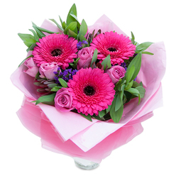 A lively, thoughtfully balanced bouquet centred on three bold fuchsia Gerbera daisies, their voluminous petals radiating outward from dark, defined centres that anchor the composition with confident contrast. Encircling these focal blooms are several lavender-pink roses in a tender, dusty-rosé shade, each bloom showing the tight, layered curls of fresh buds ready to open further. Between the roses and gerberas, clusters of tiny purple statice provide an airy texture and a cool, complementary note that lets the pinks sing without overwhelming the eye. Lush green foliage-broad leaves suggestive of salal or eucalyptus-creates a verdant frame, adding depth and a gentle, dewy freshness to the overall silhouette. The arrangement is wrapped for gifting in soft pale-pink tissue that cushions the stems, then layered with a sheer, slightly crinkled fuchsia-pink cellophane which adds a faint shimmer and a cohesive colour story; the wrapping is neatly folded and tied to indicate careful, artisan handling. The top rim of a clear glass vase is visible at the bottom, signalling immediate readiness for display. Close your eyes and imagine the soft perfume of roses mingling with the clean, green scent of foliage-an aroma that feels both tender and invigorating. Ideal for celebrating birthdays, Mother's Day, or marking an anniversary, this bouquet is presented with the kind of local care you'd expect for same-day delivery around Queensbury, Kingsbury, and nearby Wembley.