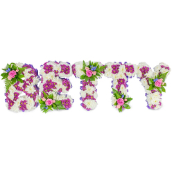 Arranged with the kind of care familiar to Queensbury families, this floral name tribute spells out BETTY in strong, freestanding capitals, photographed against a clean white background so the craftsmanship is the focus. The main mass of each letter is formed from tightly clustered, soft white chrysanthemums that create a plush, cushion-like texture and a calm, neutral ground for contrasting colours. Within and outlining the letter forms, clusters of vibrant purple and magenta chrysanthemums trace patterns and define the shapes, their deeper tones bringing visual depth and a respectful vibrancy. A slim purple ribbon wraps the sides of each letter, providing a refined edge and quiet harmony across the piece. Delicate mini-bouquets are placed atop key letters-the top left of the B, the center of the first T, and the upper right of the Y-composed of blush-pink roses in various stages of opening, tiny blue accent flowers that might be delphiniums or cornflowers, and feathery green foliage such as fern fronds. The roses add a gentle fragrance that lifts the more neutral chrysanthemum scent, while the blue specks feel like tender, hopeful details. Photographed in soft light, the arrangement reads as both dignified and comforting, a fitting tribute for a funeral service or celebration of life in Queensbury, near Kingsbury and Wembley. Prepared with local attention to detail by Flower Delivery Queensbury, it conveys personal farewell, respect and the quiet beauty families often choose to honour a loved one.