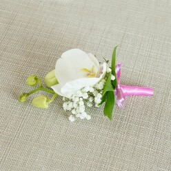 A single, immaculate white orchid boutonniere rests quietly on a light beige, linen-textured fabric, the weave of the cloth echoing the relaxed, homely feel of Queensbury's residential streets. The Phalaenopsis orchid at the centre is generously petalled, its broad white wings curving gently outward like folded silk. In its heart, a soft flush of yellow and the faintest whisper of blush pink draw the eye inward, adding a subtle depth of colour that feels intimate and refined. Surrounding the orchid's base, clusters of delicate white baby's breath form a soft, clouded fringe, each tiny bloom creating a misty, romantic halo that lightens the whole composition. A few slim, fresh green leaves slip from behind the flowers, giving a natural frame and a lively contrast to the pale tones, as if just gathered from a careful hand-selection at a local Queensbury market. To one side, several pale, lime-green orchid buds cling to a slender stem, still tightly closed and full of promise, like the careful anticipation before a wedding ceremony at a nearby registry office or hotel. The stems are neatly bound in a band of bright fuchsia satin ribbon, tied into a compact bow that glows with a glossy sheen. This striking pop of colour feels playful yet elegant, bringing a joyful edge to the otherwise gentle palette. Diffused, even lighting glides over the scene, picking out the smooth orchid petals, the airy texture of the baby's breath, and the fine threads of the linen beneath. The overall impression is of quiet celebration and meticulous craftsmanship, an understated floral accent ready to pin on for a Queensbury wedding, prom night, or cherished family event.
