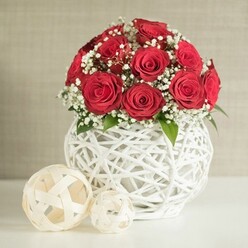 A lush dome of crimson roses rises from an artful white vase, creating a wedding-worthy table arrangement that feels at once classic and modern, like a perfectly styled reception near Queensbury's tree-lined streets. The roses, around a dozen to fifteen in number, are fully open yet still tight at the centre, their rich red petals forming deep spirals that suggest passion and devotion. Between them, airy tufts of white gypsophila float like miniature clouds, softening the intensity of the red and evoking bridal lace. Here and there, small green leaves peek through, adding a whisper of foliage that reinforces the impression of freshly arranged stems, prepared the same day for local delivery around Kingsbury and Queensbury. The bouquet sits in a round, bulbous vase crafted from interlaced white strands, an intricate open-weave design that lets light pass through, casting gentle shadows on the white tabletop below. The vase has a sculptural presence, reminiscent of handwoven decor pieces you might see in modern apartments off Streatfield Road, and it partners beautifully with the romantic roses. To the left of the vase, two woven decorative balls in soft cream tones echo the same lattice motif; the larger sphere sits slightly forward, with the smaller one tucked just behind it, giving the scene balance and a relaxed, contemporary feel. The background is a softly textured, neutral wall, unobtrusive yet warm, ensuring that all attention stays on the interplay of red, white, and subtle green. The overall effect is of refined elegance, perfect for wedding table arrangements, Valentine's dinners at home, or intimate celebrations in and around Queensbury.