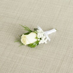 Presented on a softly textured, light beige fabric that looks like finely woven linen, a classic white rose boutonniere is arranged at a graceful diagonal, ready to be pinned to a lapel for a wedding in Queensbury. At the heart of the design is a single rosebud, its creamy ivory petals smooth and velvety, curled in tight spirals that suggest it has just opened that morning. The bloom is neither fully open nor closed, capturing a moment of quiet anticipation much like the calm before guests gather at local venues near Queensbury's high street. Nestled to one side and slightly behind the rose are airy clusters of baby's breath, their countless tiny white flowers forming light, cloud-like puffs that give the arrangement a romantic, dreamlike softness. Slender green leaves and a few feathery fronds of foliage cradle the rose, their deep green tones and gentle curves offering structure and contrast against both the white petals and the neutral background. The stems are carefully gathered and wrapped in a smooth white satin ribbon, which gleams gently under the soft light and is finished with a small, tidy bow placed just beneath the rose head, speaking to the florist's meticulous craftsmanship. The lighting is diffused and even, highlighting the textures of petal, ribbon, and fabric without harsh shadows, and allowing the creamy whites and fresh greens to appear natural and true. The entire boutonniere radiates understated sophistication and tenderness, an ideal finishing touch for a groom, best man, or guest attending a formal celebration or wedding ceremony anywhere across Queensbury and the surrounding neighbourhoods.