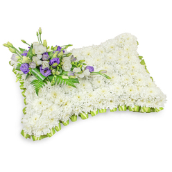A deeply respectful floral pillow, each inch covered with hundreds of pure white chrysanthemum heads, gently packed to create a smooth, pillowy surface that reads as peace itself when set against a bright white background. The chrysanthemums' rounded faces and subtle, leafy undersides form a tactile carpet that suggests softness and solemnity; their muted, green-tinged fragrance would be a delicate presence in a chapel or graveside service. The pillow's edge is outlined in a soft, light-green satin ribbon, pleated into a scalloped trim that breathes a sense of renewal into the classical white mass. Diagonally across the upper-left corner rests a modest, artful spray of accent flowers: crisp white lisianthus with layered petals, paired with rich purple lisianthus - some opening, some tightly furled - which add notes of reverence and memory. Interwoven fern fronds and darker green leaves supply natural texture and a fresh contrast, while a broader green leaf wrap neatly secures the stems and lends the cluster a hand-tied, considered finish. Photographed with gentle studio lighting and a faint drop shadow, the piece reads three-dimensionally and cleanly, reflecting the discreet craftsmanship of Flower Delivery Queensbury and local floral makers around Queensbury and Kingsbury. This pillow serves as a comforting, dignified symbol for memorial services or graveside farewells, offering a quiet, floral word of condolence when families gather to remember.