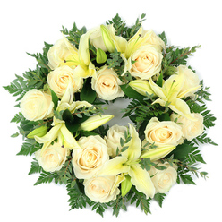 Arranged flat and photographed from directly above, this circular wreath offers a composed, compassionate presence. At its heart are about fifteen creamy white roses, their petals tightly coiled into classic rosette forms that seem to glow softly in diffuse daylight. Interspersed among them are pale yellow lilies - some petals unfurled into elegant trumpets, others still in protective buds - which add vertical motion and a hopeful sense of unfolding. The flowers rest on a luxuriant foundation of dark green foliage: delicate fern fronds provide a filigree border, while clusters of rounded, silvery-green leaves, probably eucalyptus, furnish depth and a cool counterpoint to the warm blooms. The composition is dense and balanced, the circle deliberate and continuous, a visual metaphor for remembrance and the unbroken bond of memory. There is a tactile variety here - the satiny rose petals, the smooth, waxy lily petals, and the fine softness of fern - that a careful hand would notice while arranging each stem. The scent profile seems gentle on the breeze: a soft, sweet rose note with a green, almost citrus lift from the lilies and a faint herbal coolness from the eucalyptus. Made with the respect and restraint appropriate for sympathy flowers, this wreath radiates quiet dignity and solace, a thoughtful tribute suitable for a funeral service or memorial gathering in Queensbury or nearby streets and community spaces.