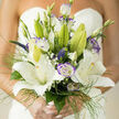 This bridal bouquet is a quiet celebration of classic form and contemporary detail, beginning with generous white lilies whose broad, velvety petals unfold with graceful confidence. Their fragrance is present but never overpowering, a soft honeyed-sweet note that mingles with fresh green undertones. Purple-tinged lisianthus punctuate the arrangement, their ruffled edges and cool mauve accents providing a refined contrast that makes the lilies' whites feel even purer. Interspersed throughout are tiny clusters of baby's breath that read like lace at a distance and reveal delicate texture up close, while narrow veronica-like sprays and fine grassy foliage introduce a sense of upward movement and airy rhythm. The bouquet is built on intentional layering: taller buds reach gently skyward, open blooms form a luminous focal core, and smaller accents trace a path around the silhouette so each stem matters. In soft daylight the petals take on a milky translucence, the violets cool and calming, and the greens vivid and fresh. As a florist at Flower Delivery Queensbury, I imagine this design arriving for a ceremony near Queensbury tube or a civil service in a local park, chosen by a bride who appreciates restrained luxury and craftsmanship. It is elegant without fuss, scented with gentle promise, and composed to be both comfortable in the hand and unforgettable in memory.