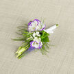 Nestled on a textured, neutral linen, this boutonniere captures a restrained springtime elegance with the careful hand of a local florist. Two white lisianthus, each edged in a whisper of violet, form the focal point; their layered, silky petals fold inward like a quiet promise. Tiny sprays of baby's breath cluster at the centre, lending an airy, almost translucent softness that offsets the deeper tones. Elongated buds and the pale stems of new growth create a sense of upward movement, while vivid grape hyacinth lends clear blue-violet spikes that punctuate the palette and add a lively counterpoint. Feathery fern fronds frame the composition, their delicate texture suggesting the cool, dappled greens of nearby parks; you can almost feel the fern's fine veins against the fingertips. A small knot of white satin ribbon binds the stems, its sheen echoing the linen's weave and the boutonniere's understated refinement. The piece is composed so it will sit gracefully on a lapel, offering tactile contrast between velvet petals and satin wrap, and releasing a barely-there, green fragrance when carried through the air. As someone who arranges blooms for couples walking past Kingsbury High Road or greeting guests near Honeypot Lane, I imagine this pinned at a ceremony in a cosy chapel or during a portrait session at a local park. Flower Delivery Queensbury prepares these with an eye for proportion and memory-the small details that make a significant day feel intimate and thoughtfully finished.