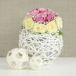 This image presents a serene and meticulously styled floral scene, filling the frame with soft colour and texture. At its heart is a low, rounded bouquet of roses in complementary shades of blush pink and buttery cream, each flower carefully placed so the colours weave together in a gentle rhythm. The roses are in full, generous bloom, their petals layered and velvety, evoking the feel of freshly opened garden roses on a still morning in Queensbury. Encircling them, delicate sprays of tiny white baby's breath float like fine lace, adding a cloud-like softness that spills just over the edge of the arrangement. A few modest green leaves peek from beneath, grounding the composition in natural foliage without stealing attention from the blooms. The bouquet rests in an unusual spherical vase made of slim white rattan-like strips, crisscrossed and intertwined into a light, open basket that suggests both craftsmanship and airiness. Through its gaps, you catch subtle glimpses of stems and shadows, giving the arrangement a sculptural dimension. On the pristine white surface around the vase, two coordinated decorative spheres made from pale, woven wood sit to one side, one larger and one smaller, echoing the orb's pattern and adding a designer's touch. The neutral beige background with its soft, horizontal, fabric-like texture frames the composition without distraction, enhancing the feeling of calm refinement. The gentle, natural-looking light makes this piece feel ready to be placed on a hallway console in a Queensbury townhouse, a cosy dining table just off the high street, or delivered as a stylish anniversary or thank-you gift by Flower Delivery Queensbury to someone who appreciates modern, understated elegance.
