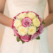 This bridal bouquet appears as a small, radiant universe of roses, the sort of design a Queensbury bride might carry as she steps out near Honeypot Lane or through the gardens by Queensbury Park. At first glance, creamy white roses dominate the composition, their petals tightly layered, satiny, and luminous, as though holding a soft glow at their centers. Intertwined among them are blush-pink and soft magenta roses, some with hints of lavender at the edges, giving the bouquet a gentle, romantic depth. The round, domed shape feels perfectly composed-no single bloom overpowering another-each rose leaning into its neighbour in a harmonious cluster. Flecks of baby's breath dot the spaces between petals, creating tiny constellations of airy white flowers that lend a dreamy, almost mist-like quality to the arrangement. From beneath the mass of roses, slender green leaves emerge in little arcs, their fresh, cool colour adding a quiet counterpoint to the warmer tones above. You can sense the texture through description alone: the velvety smoothness of each petal, the fine crispness of gypsophila, the subtle firmness of hand-tied stems wrapped together. The bouquet seems to cradle a delicate fragrance-light, floral, and clean-evoking a spring morning stroll through the neighbourhood. It radiates composure and tenderness, a bouquet designed to sit gracefully against lace or satin, to be lifted in joyful photographs outside a Queensbury venue, and to mirror the bride's inner calm and happiness on a day filled with cherished vows and new beginnings.