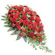 A large, traditional funeral spray in rich shades of red is carefully arranged against a pure white background, making every detail of the design easy to imagine. The flowers form a sweeping teardrop, wide and full on the upper left, then gradually narrowing to a gentle, trailing point on the lower right, like a final gesture of farewell. Deep red roses, fully opened and velvety, are scattered throughout the central area, their classic, spiralled petals giving the spray a feeling of formality and heartfelt devotion. Threaded among them are countless red carnations and smaller spray carnations, their finely ruffled petals creating layers of texture that soften the outline and make the arrangement look luxuriously full.
Around and between the blooms lies an abundance of greenery. Broad, dark green leaves provide the backbone of the spray, cupping the flowers and adding a sense of depth and stability. Among these sit smaller, silvery-green eucalyptus leaves, rounded and slightly matte, bringing a gentle, cooling contrast to the warmth of the red flowers. From the lower right edge, slender stems of lighter green foliage arch and cascade outward, adding movement and a natural, flowing finish. The overall mood is solemn but comforting, suited to rest atop a casket or at the front of a service hall in Queensbury, perhaps near the quiet residential streets off Streatfield Road. Every petal, from the velvety roses to the feathery carnations, contributes to a tribute that speaks softly of respect, remembrance, and love that lingers long after the service ends.