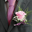 Pinned neatly to a dark, charcoal-grey lapel, this pink rose boutonniere feels like a whisper of romance captured in flowers. The central rose is a gentle, powdery pink, its petals curving inward in smooth, overlapping layers, like folded silk. It sits slightly proud of the lapel, so that its rounded form catches the soft side light, creating shadows between each petal that give it depth and dimension. Nestled around the bloom are tiny sprays of snowy gypsophila, each minuscule flower forming part of a delicate, cloud-like frame. Their crisp white colour brightens the composition and contrasts beautifully with both the rose and the darker fabric beneath. Threading through the design are slim, textural sprigs of greenery, feathery and fine, bringing a hint of the hedgerows and garden borders you might pass on an early walk through Queensbury's residential streets toward the station. The stems are bound in a lilac satin ribbon, tied smoothly and evenly, with a small, tidy tail that lies flat against the lapel. The ribbon's cool sheen subtly echoes a coordinating tie, suggesting a thoughtfully planned wedding outfit or formal ensemble. From the perspective of a Queensbury florist, this boutonniere feels perfect for a local wedding reception at a favourite restaurant or a celebration in a nearby community hall-elegant but not showy, intimate yet refined. Though the fragrance is gentle, the impression is lasting: a sense of new beginnings, of nervous smiles and held hands, conveyed through one carefully chosen rose and a handful of supporting blooms, ready for same-day delivery to homes and venues across Queensbury.