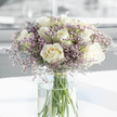 A thoughtfully composed bouquet of creamy white roses and soft purple filler flowers stands in a tall, clear glass vase, captured in a close-up that emphasises its calm, comforting presence. Each rose is in full bloom, with layers of velvety petals rolling outward in classic spirals that invite the eye to linger; their off-white, almost ivory colour carries a quiet sense of respect and remembrance. Mingling among them are clusters of tiny lavender and pale pink blossoms, probably statice, which form a gentle, cloud-like surround that lightens the arrangement and adds a subtle wash of muted colour. The filler flowers' fine, airy texture contrasts beautifully with the smooth, generous curves of the roses, giving the bouquet a soft, almost whispering quality. Through the crystal-clear cylinder of the vase, the long green stems are clearly visible, aligned with careful precision and immersed in fresh water that gleams faintly in the light. The vase rests on a pristine white surface, which reflects just a hint of the arrangement above, while the background dissolves into a blur of light, suggesting a peaceful room in Queensbury-perhaps near the quiet streets off Kenton Road-filled with natural daylight. The lighting is gentle and even, picking up the creamy highlights on the rose petals and the cool lavender tones of the surrounding florets, creating an atmosphere of stillness and gentle support. This bouquet feels like a tender tribute, the kind one might have delivered locally to offer sympathy, to stand quietly on a side table during a gathering of family and friends, and to speak of love and remembrance without the need for many words.