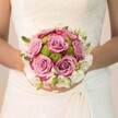 Beneath the soft glow of natural light pouring into a Queensbury ceremony hall, a bride pauses, her hands delicately supporting a standout bouquet whose beauty is both classic and heartfelt. At its center, velvety roses in soft mauve and dusty pink open wide, their ruffled petals tinged at the edges with vibrant fuchsia-a palette evoking quiet romance and joyful new beginnings. These central blooms are closely hugged by a ring of zesty lime-green pom-pom chrysanthemums, their tight, cheerful clusters echoing the vitality of neighbourhood gardens on a spring morning. Enfolding the entire composition, a gentle wreath of dainty, pure white blossoms and glossy green buds lends airiness, their presence whispering of grace and tradition. The bride's gown, adorned in intricate ivory lacework that spills softly down her arms, provides the perfect frame for this floral art. Her nails mirror the bouquet's pastel theme, polished in blush pink with a subtle spark on her ring finger, hinting at celebration and care. In this moment, local elegance and the timelessness of wedding florals converge-an ideal inspiration for brides across Queensbury dreaming of a handcrafted bouquet that reflects both community spirit and enduring love.