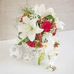 A beautifully composed floral centerpiece is arranged with the care of an experienced Queensbury florist preparing a wedding table near Stag Lane. The design sits in a round, white, open-weave basket whose intricate pattern suggests delicate wicker, allowing glimpses of stems and adding a handmade, artisanal charm. Emerging from this airy container, several large white oriental lilies command attention, their sculptural petals fully open, revealing prominent stamens tinged with pollen and subtle shadows that hint at their soft, almost velvety texture. Nestled among these lilies are rich red roses, their velvety petals tightly wound into classic spirals that speak of romance and devotion, ideal for wedding receptions or anniversary dinners across Queensbury and neighbouring Harrow. Wisps of white baby's breath thread their way through the larger blooms, bringing an ethereal lightness, while fine, feathery green foliage creates movement and depth as though a gentle breeze had just passed through. Clusters of bright red hypericum berries glint like tiny ornaments, adding small but vivid bursts of colour that subtly nod to festive gatherings. On one side of the composition, a graceful spray of white Phalaenopsis orchids cascades downward, each bloom edged delicately and centred with a hint of yellow and pink, accompanied by a few firm buds that suggest continued flowering over the coming days. Resting on the white tabletop to the left are several small, white woven spheres that mirror the basket's texture, visually anchoring the piece and giving it a contemporary, styled look. The softly textured beige background and gentle lighting keep the focus squarely on the interplay of white, red, and green, creating a mood of serene elegance and joyful celebration.