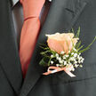 This image captures a single peachy-pink rose boutonnière carefully arranged and fastened to the left lapel of a deep charcoal suit, described with the attentive eye of an experienced Queensbury florist. The rose is the composition's heart: petals layered with soft density, edges curling outward as if opening to a quiet morning. Gentle daylight highlights the rose's velvety texture, rendering the peach tones luminous against the suit's cool grey. Sprays of tiny white gypsophila hover around the bloom like miniature clouds, while wisps of fine green foliage tuck in and trail slightly, lending a hand-crafted asymmetry that feels both natural and considered. At the base, a slim satin ribbon in a pale salmon-peach hue is tied into a restrained bow, its satin sheen echoing the thread of colour in the wearer's tie and drawing the eye.
The tie beneath shows a smooth peach hue with a faint, dark grid on a light shirt, offering a soft graphic counterpoint to the organic curves of the blossom. The charcoal jacket presents a crisp, structured canvas so the boutonnière's softness reads as intentional warmth-perfect for a wedding or refined celebration. Close enough to imagine the rose's subtle scent, the image communicates freshness: stems trimmed cleanly, foliage wired and positioned for comfort and longevity.
Rooted in local sensibility-fit for couples exchanging vows near Queensbury Broadway or greeting guests arriving at Kingsbury-the arrangement reflects careful colour coordination and the quiet expertise of a florist who knows how to translate garden notes into wearable elegance.