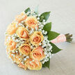 There is a classic tenderness to this hand-tied bouquet: apricot-peach roses arranged in a compact, rounded formation so each bloom nestles into the next, petals curling softly in layered spirals. Interspersed throughout is a delicate spray of baby's breath (gypsophila), its tiny white flowers forming a light, cloud-like contrast that accentuates the roses' creamier tones. Broad, deep-green leaves frame the composition, their rich colour and glossy texture adding depth and an organic structure that keeps the design grounded. The stems are bound in a textured peach ribbon trimmed with pearl pins, a tactile finish that reads both elegant and personal under gentle, diffuse light. The soft daylight highlights the creamy undertones and reveals a subtle fragrance-fresh, green-cut foliage mingling with the sweet, old-fashioned scent of roses and the whisper of gyp. As a local florist familiar with Queensbury High Road and nearby Wembley gardens, I often recommend this style for brides seeking a timeless look that photographs naturally in the soft light of a church or village hall. Available for local delivery through Flower Delivery Queensbury, the bouquet balances classic bridal romance with modern restraint: it is simple enough to carry with ease, yet detailed enough to feel treasured, offering both comfort and a gentle promise of celebration.
