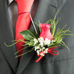 This photograph shows a finely crafted wedding boutonnière, designed with the care and precision you might expect from a trusted neighbourhood florist in Queensbury. The focal point is a beautifully formed hot pink rose, its petals arranged in tight, velvety layers that open outward like a slow, deliberate breath. The shade is both romantic and modern, a vivid note of colour against the dark lapel of a well-cut suit. Surrounding the rose, tiny white blossoms of baby's breath hover like a ring of delicate snowflakes, their clustered heads adding an airy softness and a sense of purity to the design. Slender, looping strands of ornamental grass arc around the central bloom, lending the piece movement and a subtle sense of drama, reminiscent of the gentle sway of greenery in gardens off Church Lane and around Queensbury Circle. Behind the composition, a glossy deep green leaf anchors the boutonnière, creating structure and a natural frame that makes the rose stand out even more. At the base, a neat red satin ribbon is tied with care, its small bow signalling the handcrafted nature of the arrangement and catching the light with a quiet sheen. The contrast between the bright florals and the dark, textured fabric of the suit adds sophistication, making this boutonnière perfect for weddings, proms, or anniversary celebrations held in and around Queensbury. Every element-from colour to texture-comes together to express tenderness, commitment, and a love of beautiful details on a day meant to be remembered.
