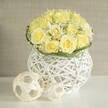 A contemporary yet romantic wedding centerpiece is shown as if arranged for a stylish reception in Queensbury, perhaps in a boutique venue not far from Queensbury Underground Station. At its heart is a compact, dome-shaped bouquet of pale butter-yellow roses, so closely clustered that their velvety petals form a seamless mound of colour. Each rose is fully open, revealing layered spirals that hint at a gentle, sweet fragrance. Between the roses, airy sprays of white baby's breath thread through the design, their tiny round blossoms creating a soft halo effect, like a fine mist drifting over a garden at dawn. The interplay of creamy yellow and crisp white produces a light, uplifting palette that feels both elegant and approachable.
A narrow collar of rich green foliage hugs the base of the bouquet, its sleek leaves arranged with precision to create a defined edge where the flowers meet the container. This container is a sculptural white woven vase, rounded and slightly cinched at the waist, made from slender strands that interlace to form an open, basket-like lattice. The structure reads as modern and airy, allowing glimpses of the stems within and casting delicate, crisscrossing shadows onto the smooth white table surface below. Placed beside the vase are two decorative spheres crafted from pale, rattan-style strips, their open geometric construction echoing the vase's woven texture and giving the scene a harmonious, curated look.
Behind the arrangement, a softly blurred wall in warm beige tones provides a calm, neutral backdrop, similar to the soothing interiors of many Queensbury flats and family homes. The image conveys a sense of gentle celebration and polished minimalism, ideal for wedding tables, anniversary lunches, or intimate gatherings where Queensbury hosts want flowers that feel fresh, local, and quietly refined.