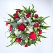 Viewed directly from above against a clean white backdrop, this luxurious round bouquet looks as though it has just been set down on a florist's bench in Queensbury. At its centre, velvety crimson red roses form a rich heart, some still in tight bud, others just beginning to unfurl their layered petals, suggesting unfolding romance and quiet elegance. Clustered around them are deep fuchsia and ruby red carnations, their frilled, ruffled heads adding a sense of movement and plush texture, like festive confetti caught mid-dance. Nestled between the bolder blooms are small white spray chrysanthemums, daisy-like and cheerful, their bright centres and crisp petals bringing light and balance to the composition. Scattered clouds of tiny white gypsophila, or baby's breath, float through the bouquet, softening every edge with a misty, almost shimmering effect, as if the flowers are wrapped in morning haze above Queensbury Park. Lush greenery frames the display in a generous halo. Strong, pointed dark green leaves, likely from lilies or alstroemeria, radiate outward to give structure, while rounded, silvery-green eucalyptus leaves weave between stems, offering a cool, contemporary touch and a hint of soothing scent. The flowers sit densely together, forming a full, generous dome that feels both modern and timeless. The natural daylight in the photograph brings out the true reds, whites, and greens, making each texture-from rose velvet to chrysanthemum crispness-easy to imagine under your fingertips. This is the kind of bouquet that might be delivered to a doorstep off Kenton Road to celebrate an anniversary, mark a birthday, or simply send a vivid embrace of love and good wishes across Queensbury.