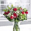 A rich, romantic bouquet of deep crimson red roses and ruffled carnations stands proudly in a clear glass vase on a bright white tabletop, bathed in gentle daylight from a large, softly blurred window behind. Each velvety rose, some fully open and others still in tight bud, shows off layered petals that seem almost touchable, while the carnations in the same bold red add volume and a nostalgic, traditional charm. Between the reds, pristine white alstroemeria-Peruvian lilies-peek through, some still closed like little green-tipped torches, others just beginning to open, revealing their elegant, trumpet-shaped blooms. Tiny white gypsophila, or baby's breath, is scattered throughout the arrangement, creating a cloudlike mist that softens the stronger lines and colours, giving the bouquet a dreamy, romantic halo. Lush green foliage frames the flowers: broad, dark green leaves form a structured collar at the base, while slimmer, lighter green stems weave upward, adding movement and contrast. Through the clear glass, you can see the neatly cut stems fanning into fresh water, underscoring how newly prepared and vibrant this design feels. The white surface beneath the vase reflects a soft glow back into the arrangement, heightening the reds and whites against the almost grayscale view outside the window. As a florist in Queensbury, this is the kind of bouquet I'd recommend for heartfelt anniversaries, romantic surprises, or milestone celebrations at home, a piece that could just as easily brighten a dining table near Queensbury Circle as it could grace a living room overlooking one of the quiet residential streets off Kenton Road, bringing a sense of joy, tenderness, and thoughtful elegance into the space.