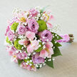 On a softly textured, light grey linen surface, a lush bridal bouquet lies gently on its side, as if just set down between photographs on a Queensbury wedding morning. The flowers form a rounded, compact shape, with velvety lavender roses taking centre stage, their petals layered so densely that they appear almost sculpted, each bloom cradling a hint of shadow at its heart. Woven among them, pink alstroemeria stretch their slender petals outward, washed in blush pink with touches of creamy white and sunlit yellow near the throat, marked by subtle freckles that give them a natural, garden-grown charm. Small orbs of bright chartreuse button chrysanthemums punctuate the design like playful notes of colour, their tightly clustered petals creating a modern, textural counterpoint to the softness of the roses. Tiny white gypsophila sprigs disperse through the bouquet, adding a misty, cloud-like lightness that softens the overall silhouette and brightens the pastel tones. The stems are firmly bound at the base, wrapped in a wide pink satin ribbon that gleams under gentle light, then spiralled with a line of pearly beads, reminiscent of bridal accessories seen in Queensbury's local boutiques. Below this polished handle, the natural stems peek out, slightly uneven and organic, reinforcing the hand-crafted, personal feel. The composition suggests romance and optimism, suitable not only for a ceremony but also for engagement dinners or anniversary celebrations around Queensbury. It is an arrangement that feels both timeless and current, as if created by a florist who knows the rhythm of local love stories and the subtle elegance that couples here often seek.