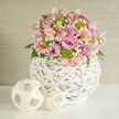 This image captures a finely crafted floral centerpiece that feels both romantic and modern, as if it has just been styled in a boutique studio close to Queensbury station. The bouquet forms a soft dome of colour, led by generous roses in shades of lilac, soft mauve, and rosy pink, their petals layered like silk and opening just enough to reveal their intricate hearts. Nestled among them are pale pink alstroemeria with speckled throats, the kind of detail you might notice when pausing over coffee in a Queensbury café, their star-shaped blooms adding movement and delicacy. Bright lime green button chrysanthemums provide a striking contrast, their perfectly rounded forms echoing the overall shape of the bouquet and lending a fresh, contemporary edge. Threaded throughout are tiny tufts of white baby's breath, creating a misty veil that lightens the design, while dark green foliage offers subtle depth beneath the blossoms. The flowers are gathered into a spherical white vase made from finely woven wicker or rattan, its open, basket-like structure giving the arrangement a floating quality and a touch of Scandinavian-inspired style. On the gleaming white surface beside it sit two smaller woven orbs in a pale, natural wood tone, mirroring the texture of the vase and adding an extra layer of visual interest. The softly textured, neutral wall behind allows the gentle pastels and greens to stand out clearly, creating a serene, uplifting mood perfect for a wedding collection, bridal shower display, or sophisticated Queensbury home, with same-day delivery easily imagined from a nearby local florist.