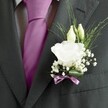 A close-up photograph captures the left lapel of a man's dark charcoal grey suit, the fabric a fine wool with a subtle woven texture that looks crisp and freshly pressed, as if readied for a wedding at Queensbury's parish church. At the centre of the image sits a carefully crafted buttonhole: a single white lisianthus bloom, fully open, its many soft, ruffled petals forming a rounded, cloud-like flower. The petals are creamy rather than stark white, with gentle shadows that hint at a velvety touch. Just above the main blossom, two slender ivory buds lean in, still closed and elegant, suggesting flowers about to unfurl later in the day. Surrounding them is a light spray of baby's breath, each tiny Gypsophila flower a miniature white dot that softens the arrangement and gives it a romantic, misty halo. Fine strands of fresh greenery arc outward, thin and slightly curved, adding texture and a sense of airy movement against the structured lines of the suit. The base of the boutonnière is neatly wrapped in a narrow mauve ribbon, tied into a delicate bow that mirrors the rich magenta or deep fuchsia silk tie resting between the jacket's lapels. The tie is smooth and lustrous, perfectly knotted against a grey-and-white patterned dress shirt, creating a refined balance of colour and pattern. The overall mood is one of quiet anticipation and sincere celebration, reminiscent of a groom pausing outside one of Queensbury's leafy streets before the ceremony begins, his flowers as thoughtfully chosen as his promises. Crafted with a florist's precise hand, the arrangement feels fresh, cool, and lightly scented, an intimate detail that completes a formal, emotionally charged moment.