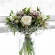 In the centre of the image, a graceful sympathy bouquet rises from a clear glass vase, its gentle whites and soft purples capturing the stillness of a quiet moment in Queensbury. Dominating the design are open white roses, their velvety petals arranged in perfect spirals that feel both tender and composed, like a whispered farewell. Between them drifts a generous scattering of baby's breath, each tiny white flower contributing to a mist-like cloud that softens the outlines of the larger blooms. Threaded throughout, slender stems of alstroemeria show buds and half-open flowers in muted shades of lavender, pale purple, and rose-lilac, with fine, natural striping that adds interest without disturbing the bouquet's calm character. Around the base, deep green tropical-style leaves are folded and curved into a smooth band, cradling the heads of the flowers, while thinner green leaves weave upward, lending subtle movement and contrast. Below, the straight green stems are clearly visible through the vase, submerged in clear water and gathered neatly with a narrow binding so the arrangement keeps its rounded, hand-tied profile. The background is bright yet softly blurred, as if the bouquet were placed near a sunlit window overlooking a quiet side street near Queensbury Circle, creating an atmosphere of peace and reflection. With its restrained palette, refined structure, and delicate details, this bouquet embodies solace and remembrance, the kind of flowers chosen to offer comfort to a grieving family or to stand thoughtfully at a memorial table in the local community.